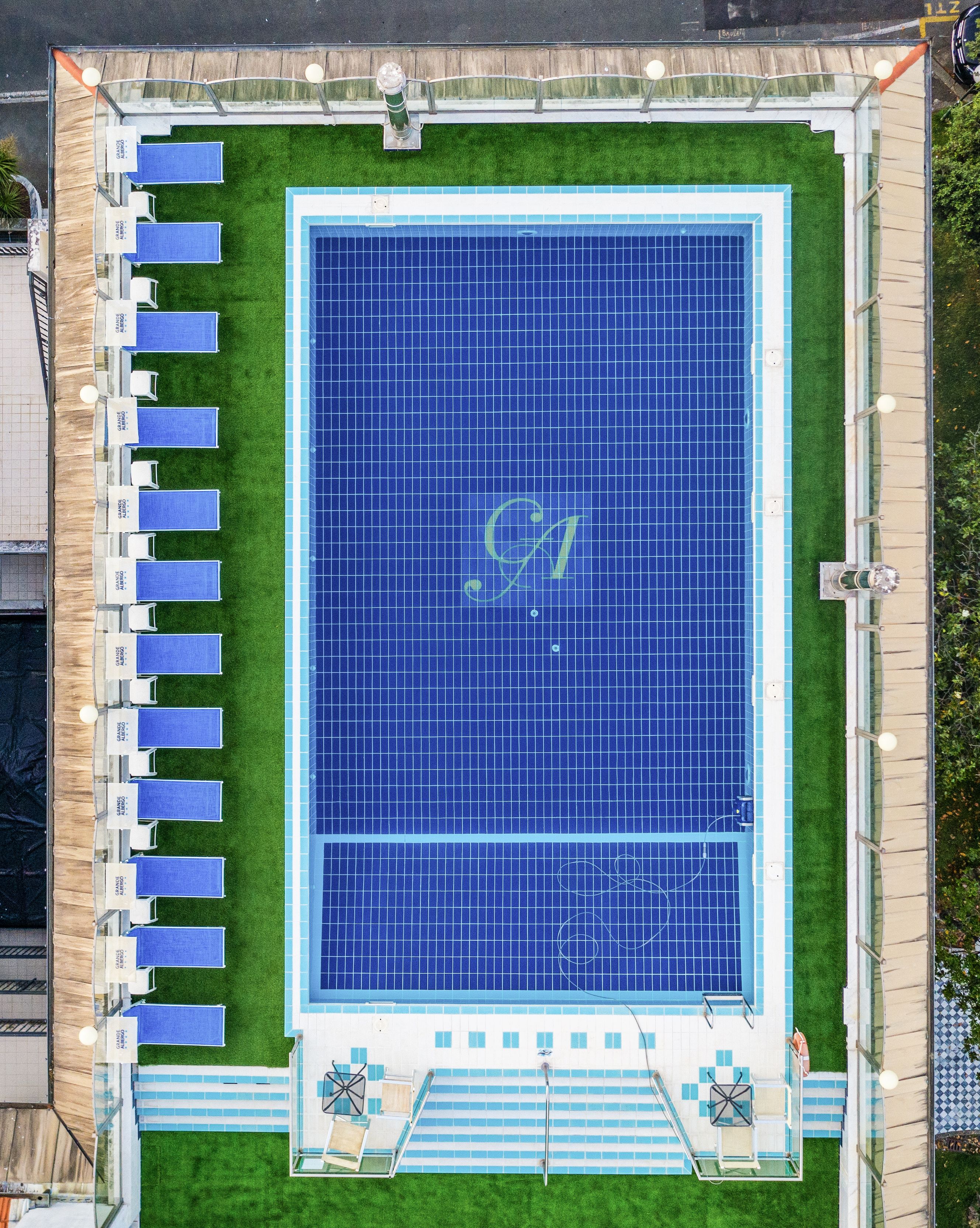 An aerial view of a swimming pool with a large 'G' logo, surrounded by a deck area with lounge chairs and a small poolside table.