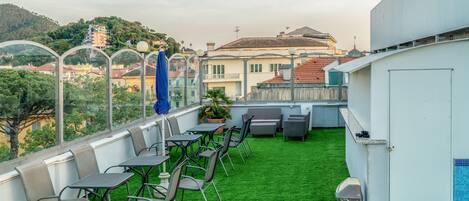 A rooftop terrace with green artificial grass, metal tables and chairs, and a view of surrounding buildings.