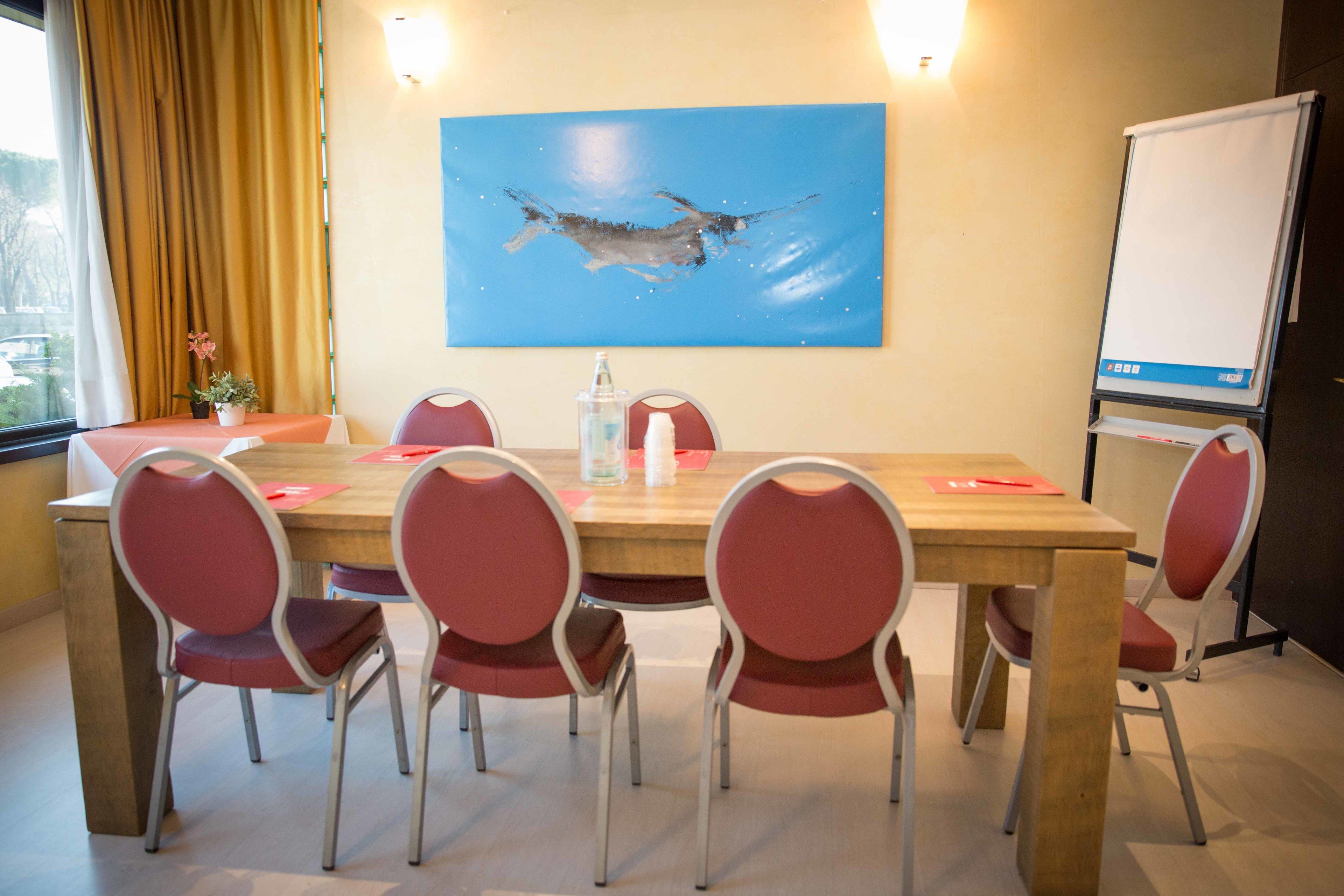 Un salón de conferencias con una mesa de madera, sillas rojas, un cuadro de un pez, una pizarra y una ventana con cortinas.