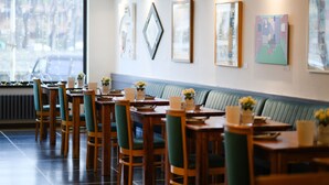 Un restaurante con mesas de madera, sillas tapizadas de verde, un espacio con bancos corridos y obras de arte en las paredes.