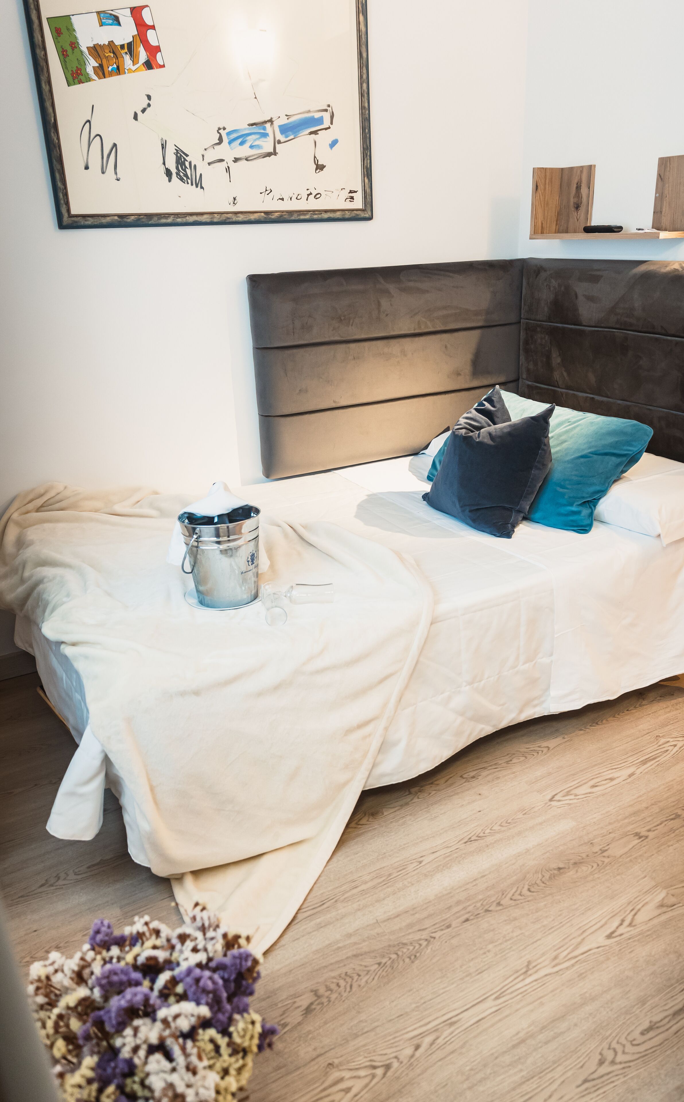 A neatly made bed with a white bedspread, a metal bucket, and a glass bottle on the floor beside it.