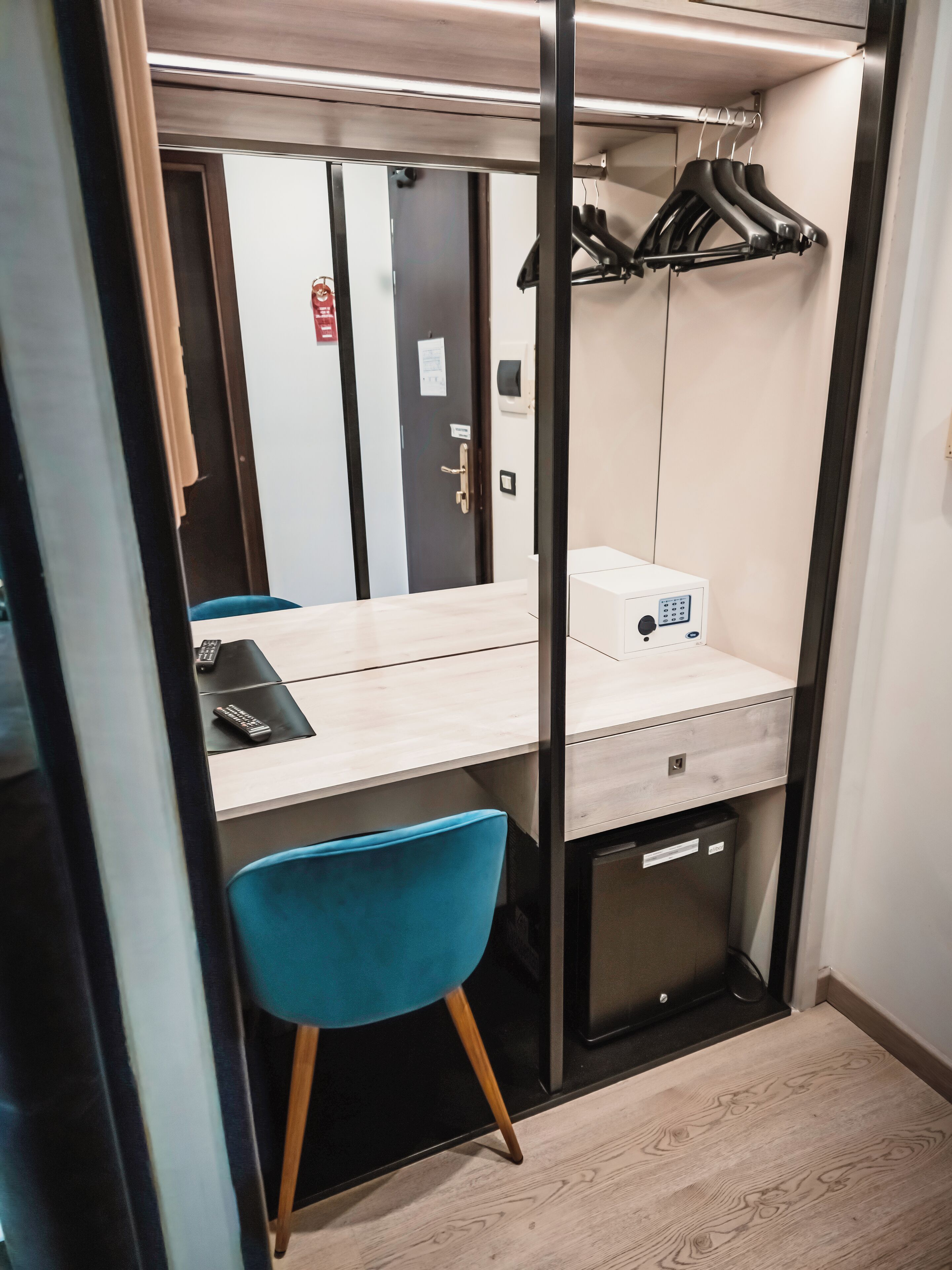 A compact hotel room with a wooden desk, a blue chair, a mirror, and a wardrobe with hanging clothes.