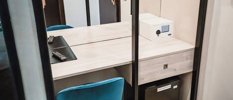 A compact hotel room with a wooden desk, a blue chair, a mirror, and a wardrobe with hanging clothes.