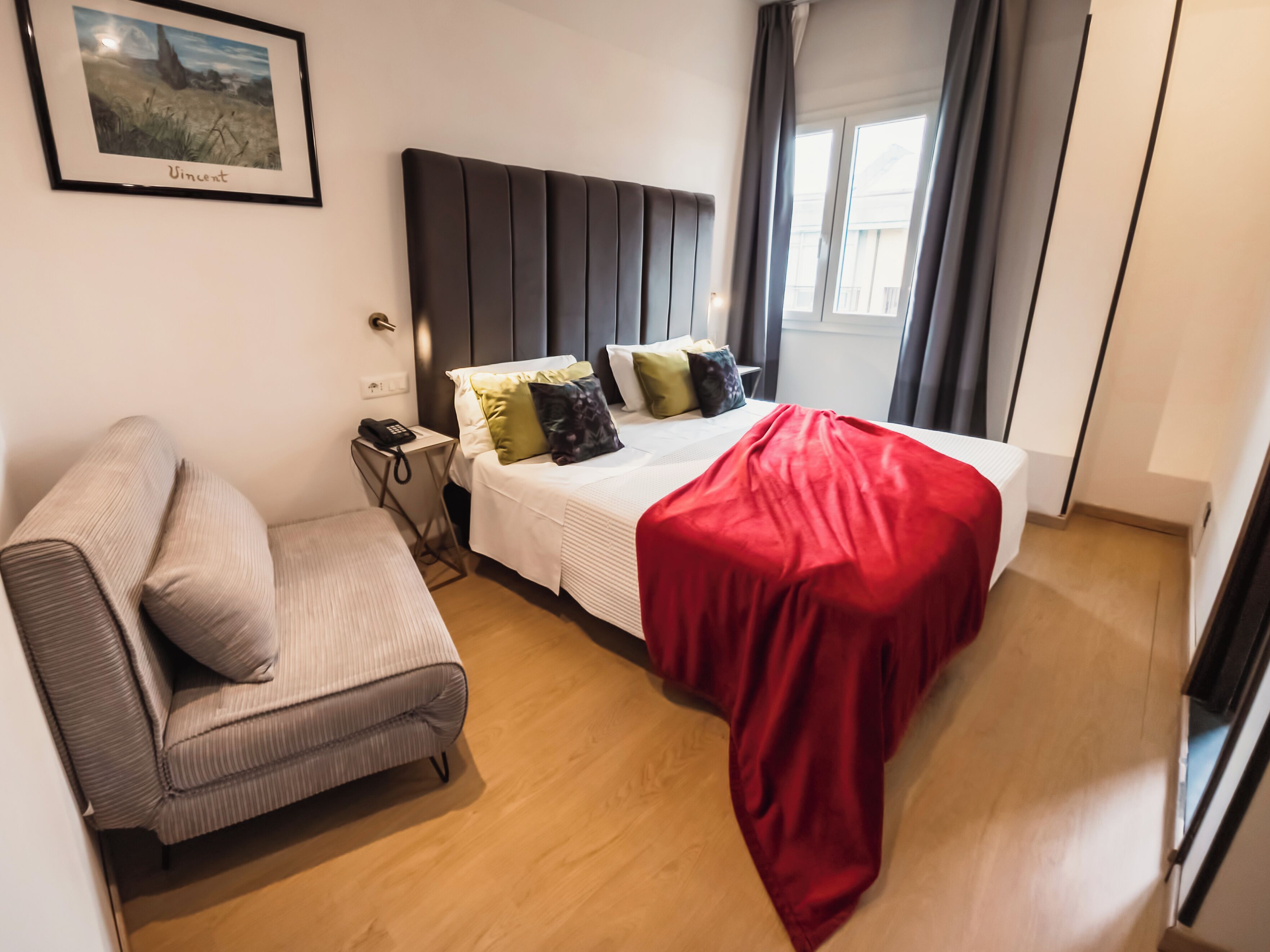 A hotel room with a bed, a sofa, a bedside table with a phone, and a window with curtains.