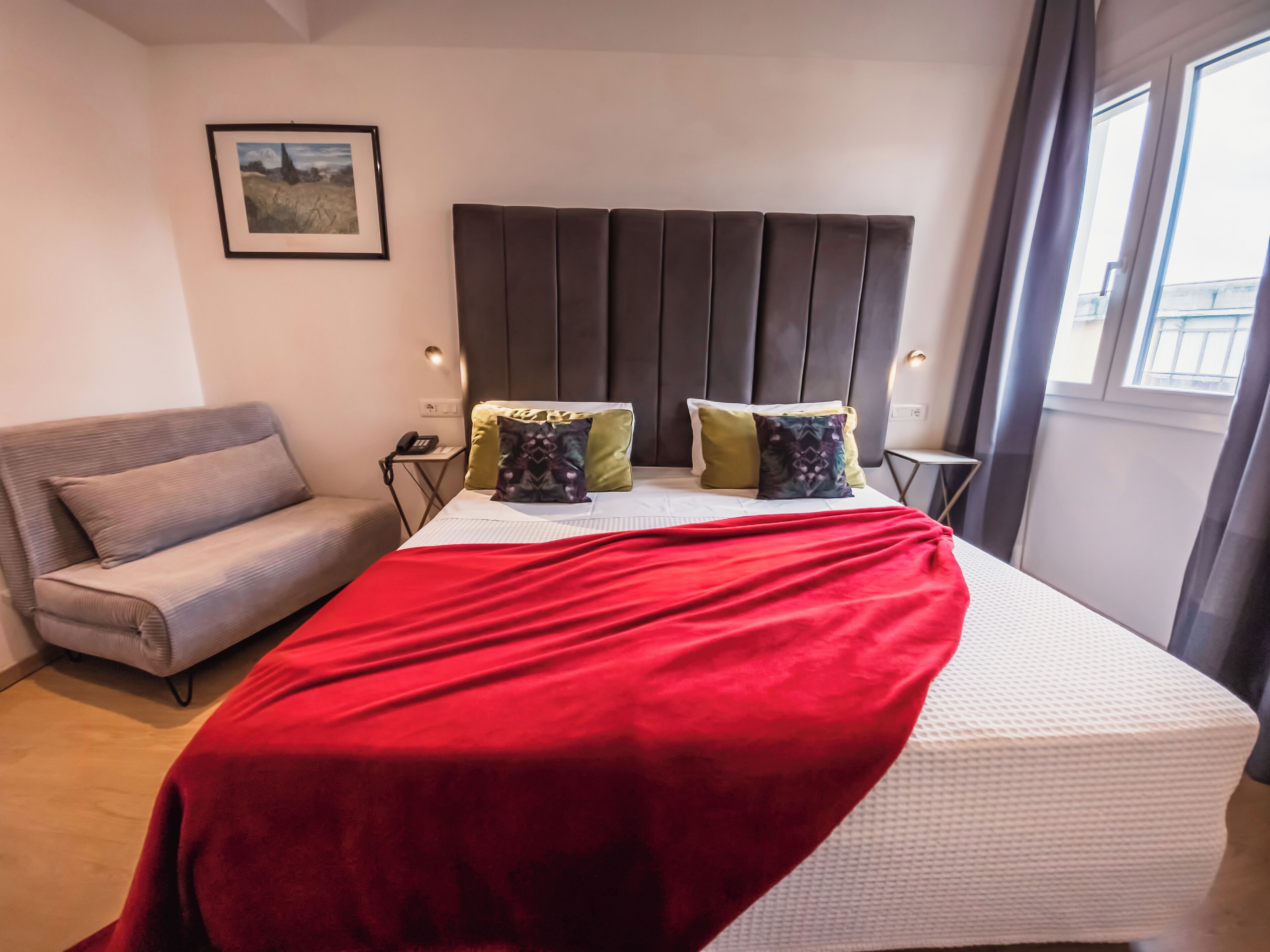 A hotel room with a bed, a sofa, a bedside table with a phone, and a framed picture on the wall.