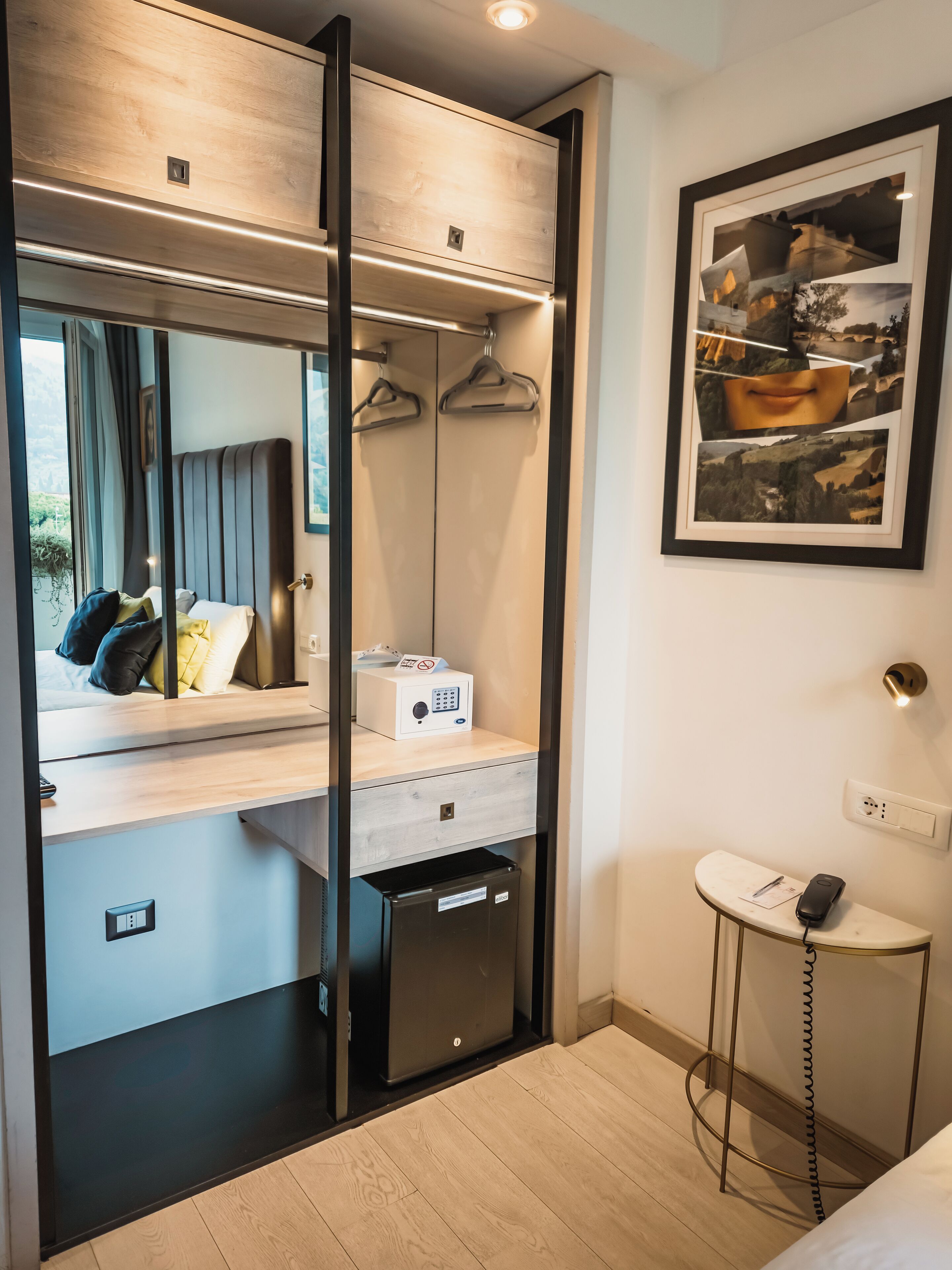 A compact room with a bed, a desk, a wardrobe, a small table with a phone, and a framed picture on the wall.