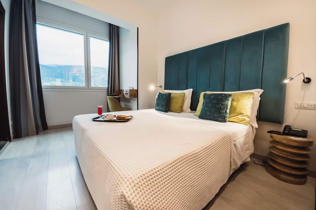 A hotel room with a large bed, a bedside table with a lamp, a view of the city through the window, and a cityscape visible outside.