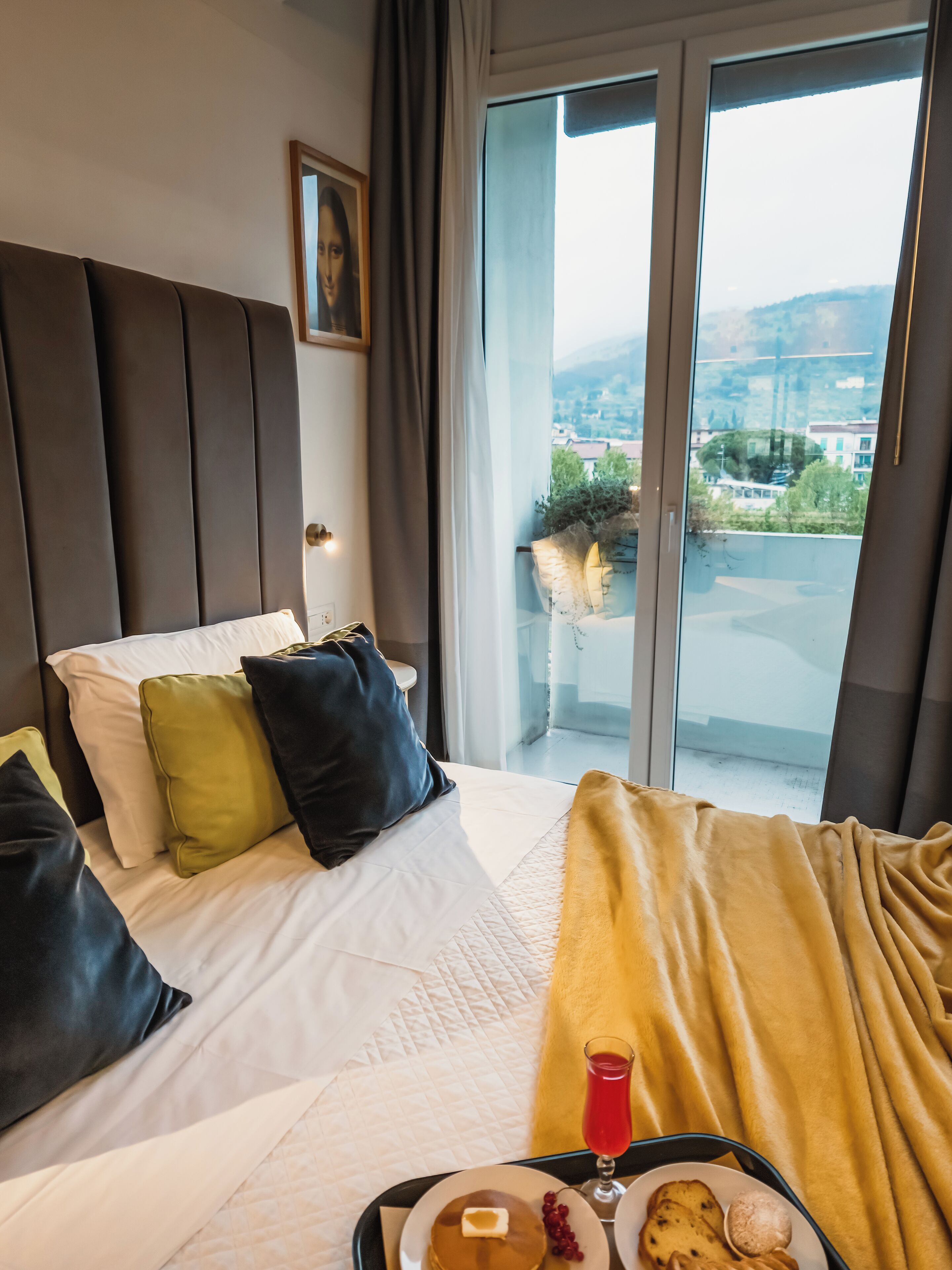 A hotel room with a bed, a bedside table with a tray of breakfast, a view of the city through the window, and a painting on the wall.