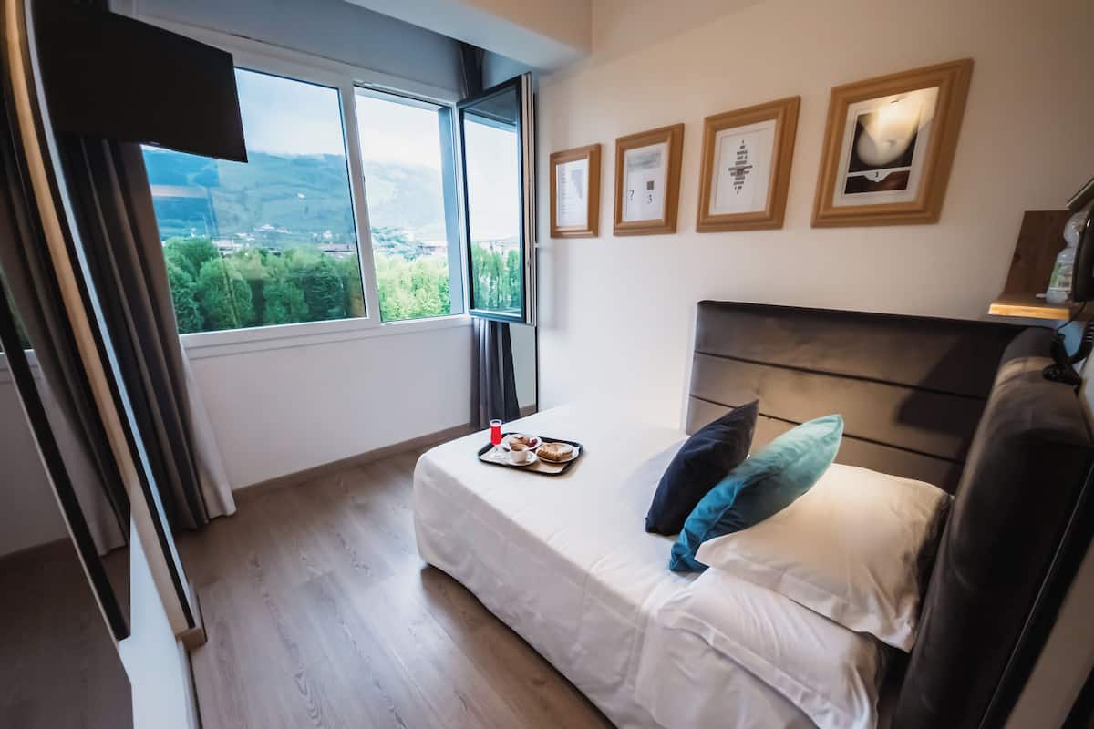 A bedroom with a bed, a window with a view of greenery, and framed pictures on the wall.