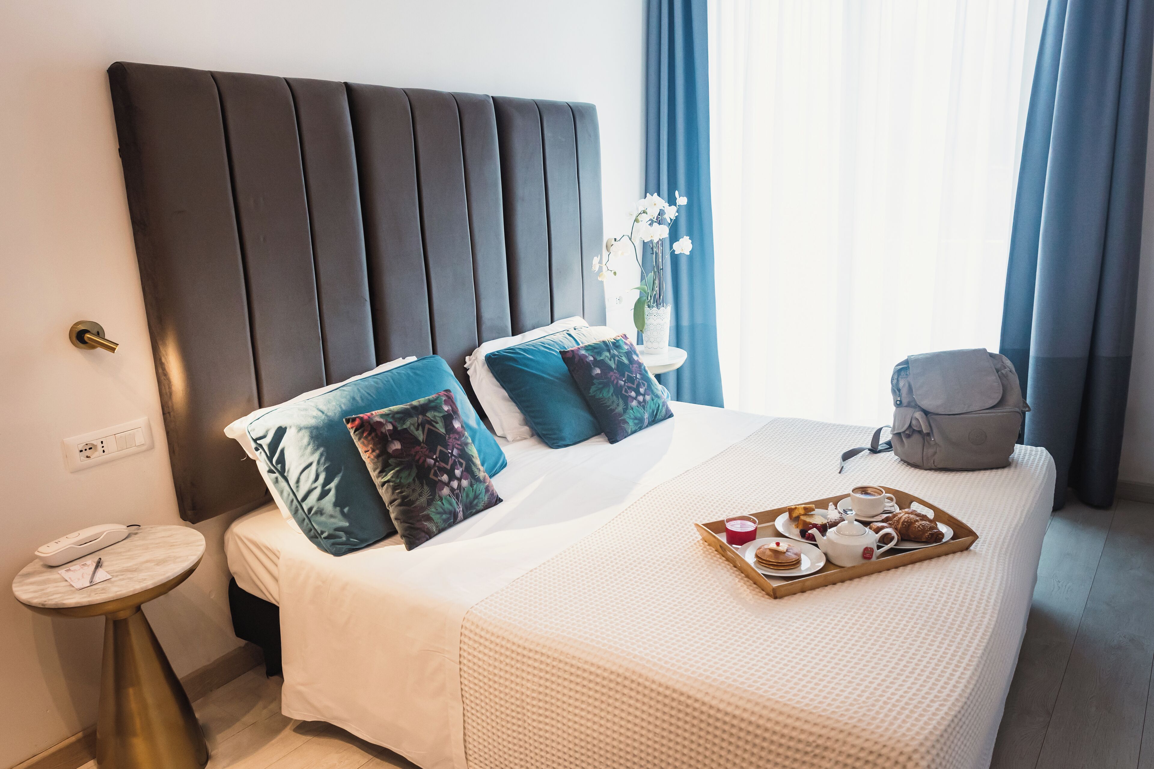 A neatly made bed with a tray of breakfast items, a bedside table with a phone, and a window with curtains.