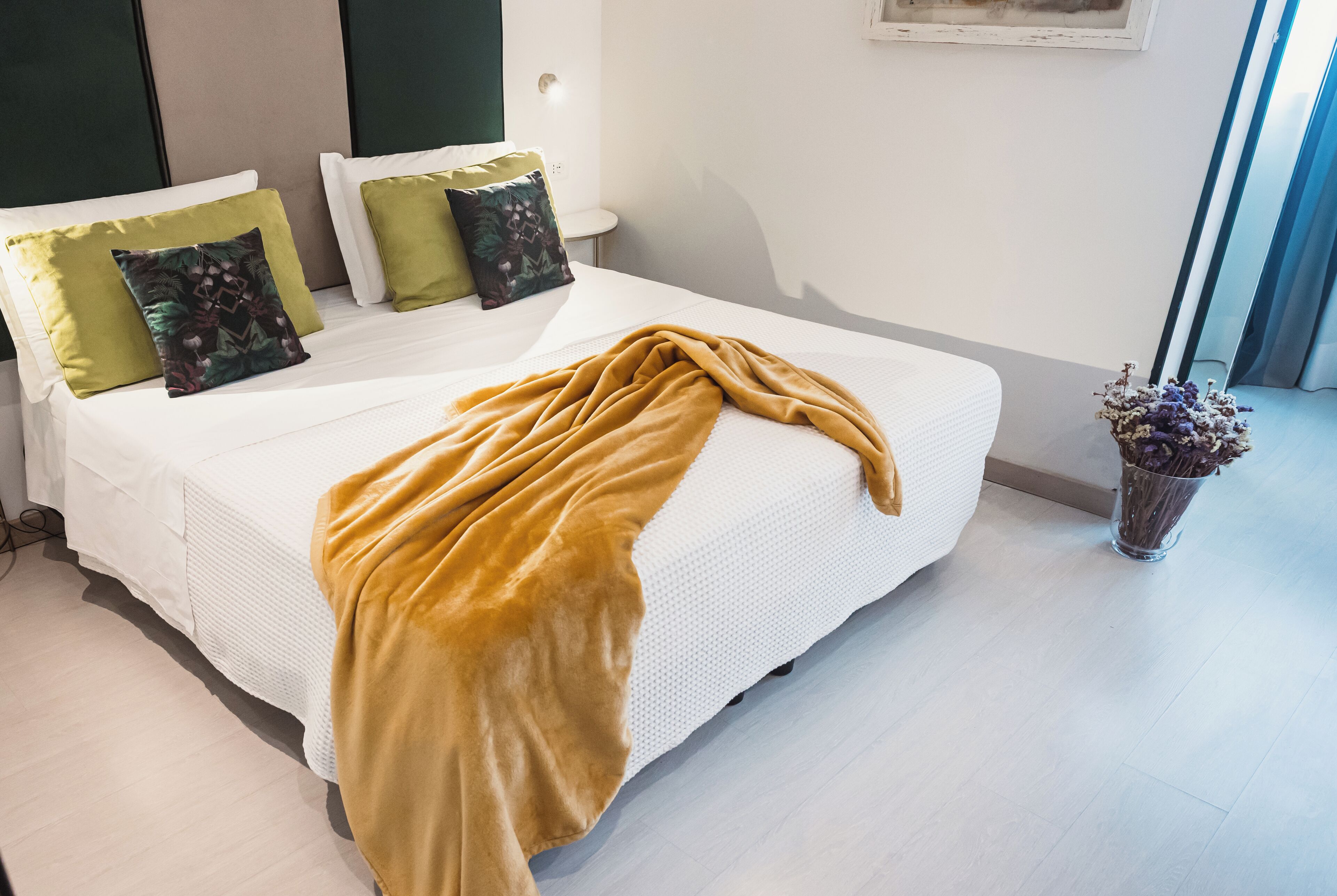 A neatly made bed with a mustard-colored blanket, white bedding, and green and patterned pillows.