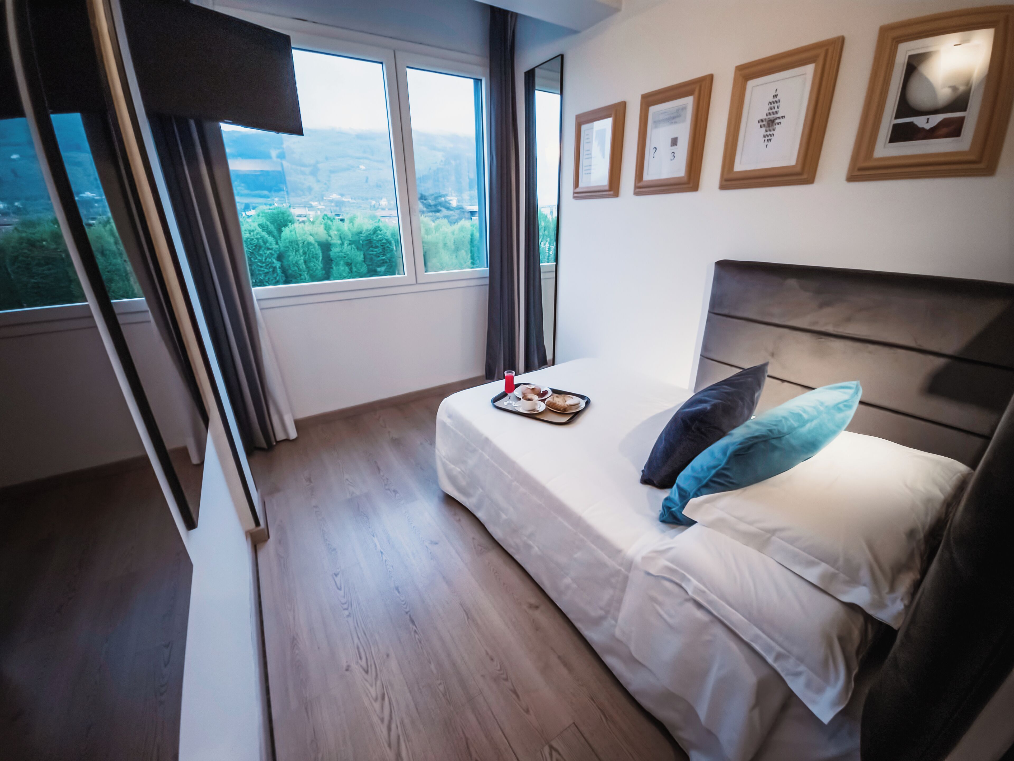 A bedroom with a bed, a tray with breakfast items, a window with a view of greenery, and framed pictures on the wall.