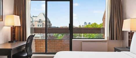 A hotel room with a bed, desk, chair, and large windows offering a city view.