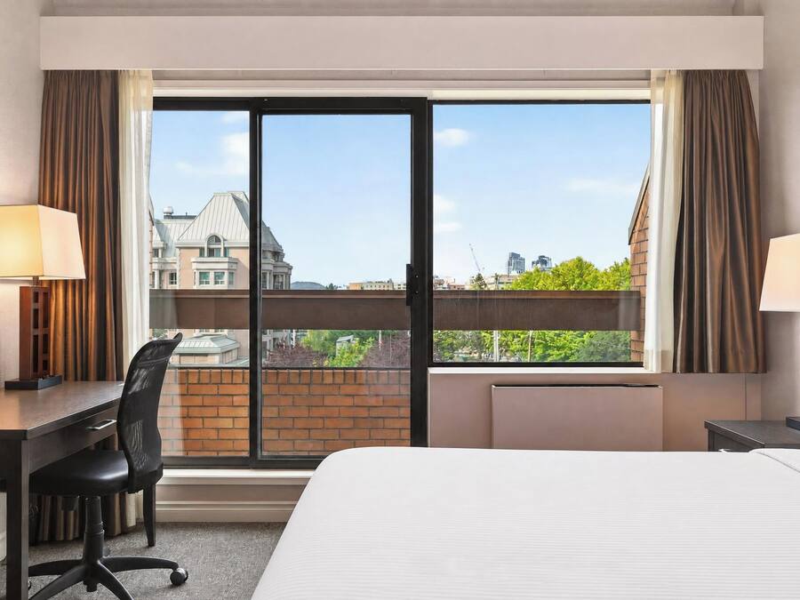 Una camera d'hotel con un letto, una scrivania, una sedia e ampie finestre che si affacciano sulla città.
