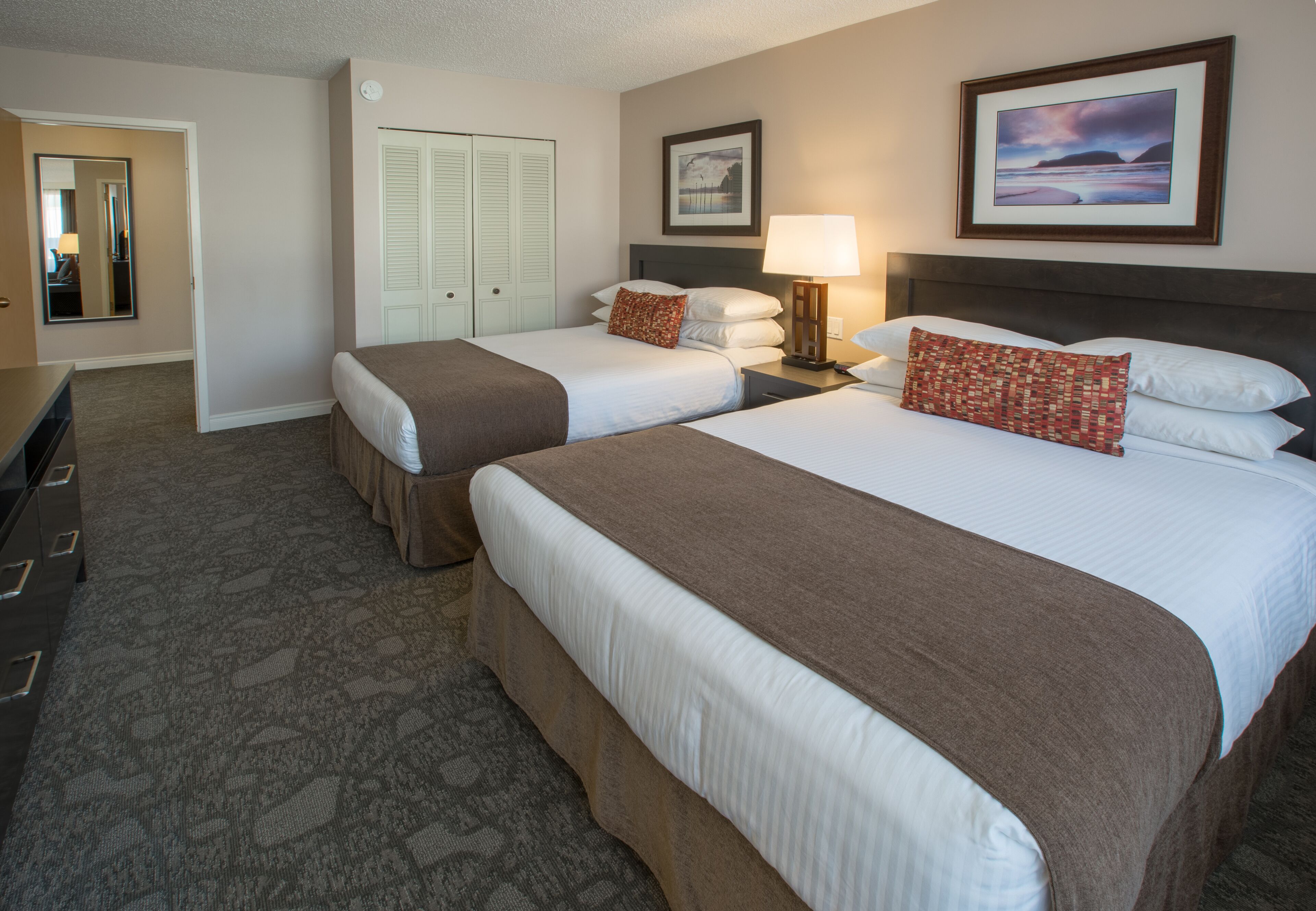 A hotel room with two beds, a dresser, a mirror, a door, and a framed picture on the wall.