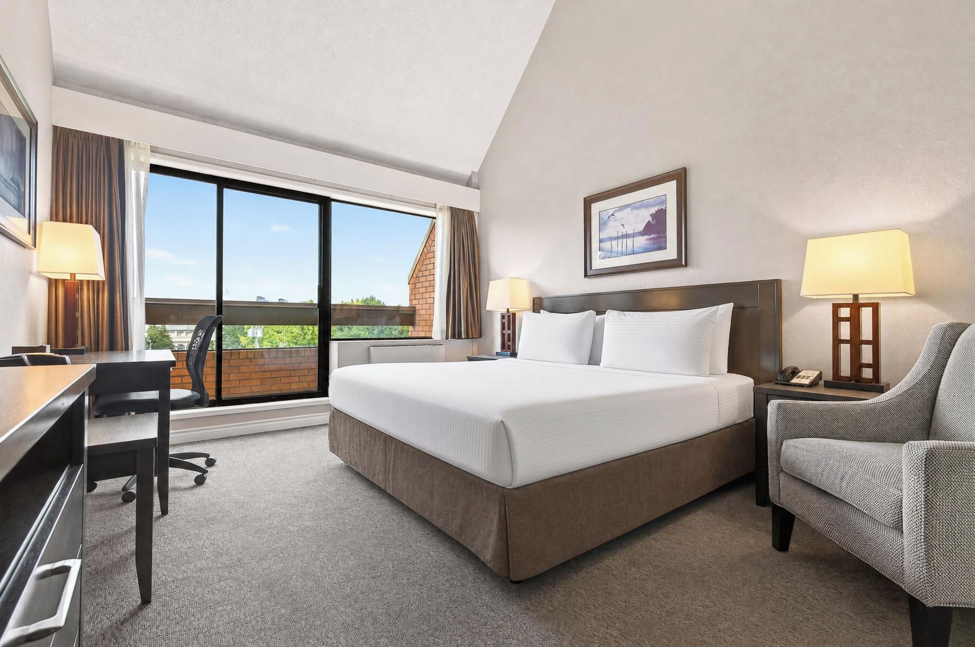 A hotel room with a large bed, a desk, a chair, a window with curtains, and a framed picture on the wall.