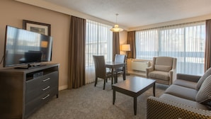 A hotel room with a flat-screen TV, a dining table with chairs, a sofa, a lamp, and large windows with curtains.