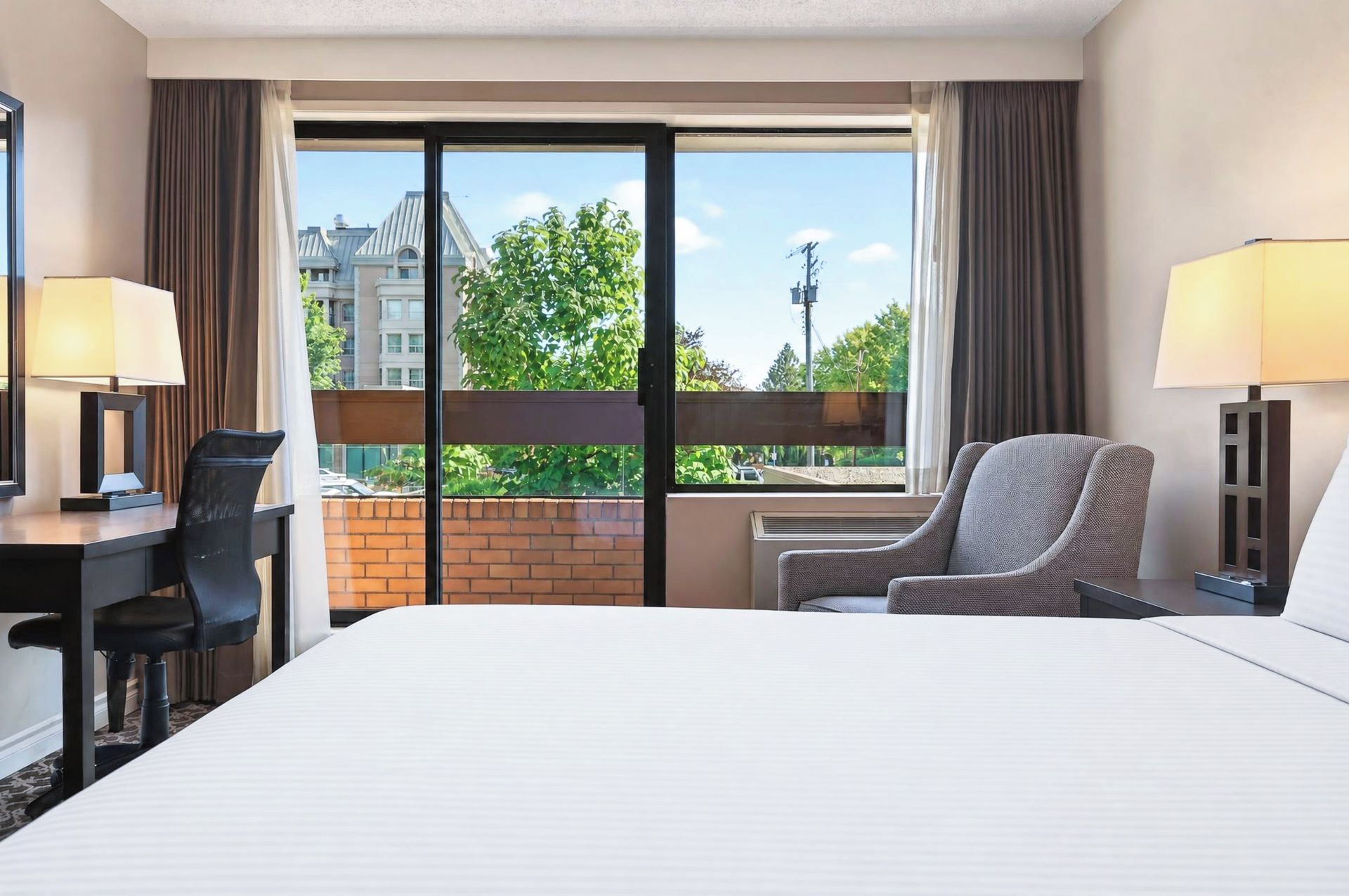 A hotel room with a bed, a desk, a chair, a window with a view of buildings and trees, and two lamps.