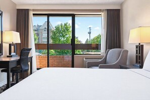 Een hotelkamer met een bed, een bureau, een stoel, een raam met uitzicht op gebouwen en bomen, en twee lampen.
