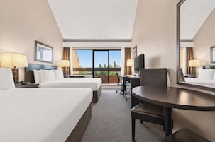 A hotel room with two beds, a desk, a chair, a television, and a large window offering a view of the outdoors.