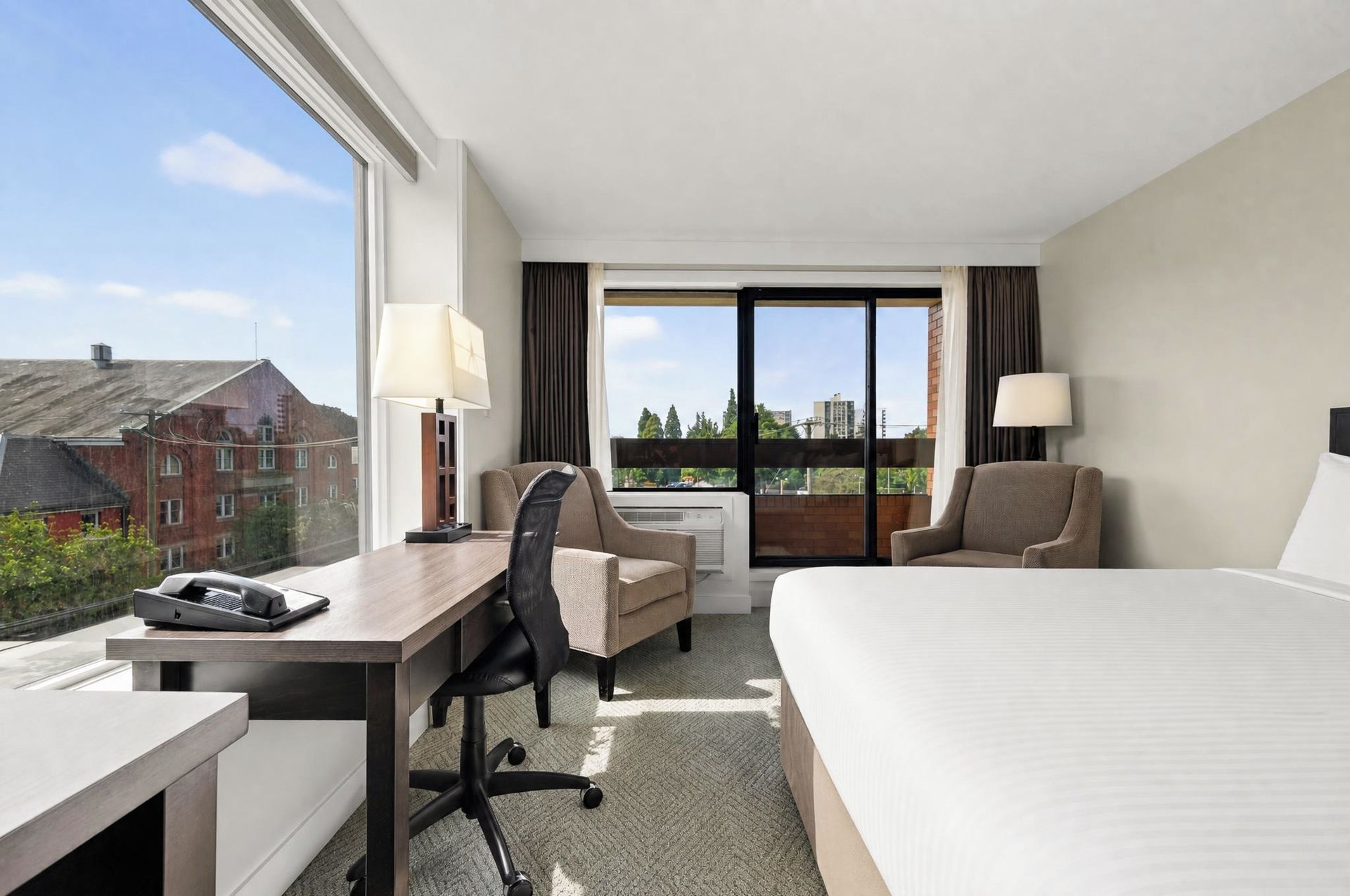 A hotel room with a bed, desk, two chairs, a window with a view of buildings, and a lamp.