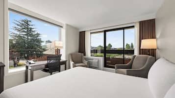 A hotel room with a large bed, a desk with a lamp, a chair, and a view of the city through large windows.