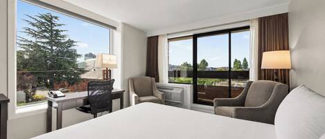 Habitación de hotel con una cama grande, un escritorio con lámpara, una silla y vistas a la ciudad a través de amplios ventanales.