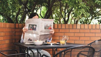 A person reading a newspaper at an outdoor table with chairs, a brick wall, and trees in the background.