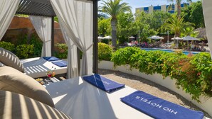 Ein sonniger Außenbereich mit Liegebereich, Pool, Palmen und Blick auf ein Gebäude im Hintergrund.