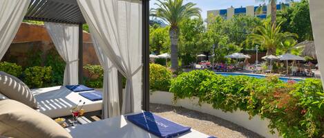 A sunlit outdoor lounge area with a pool, palm trees, and a view of a building in the background.