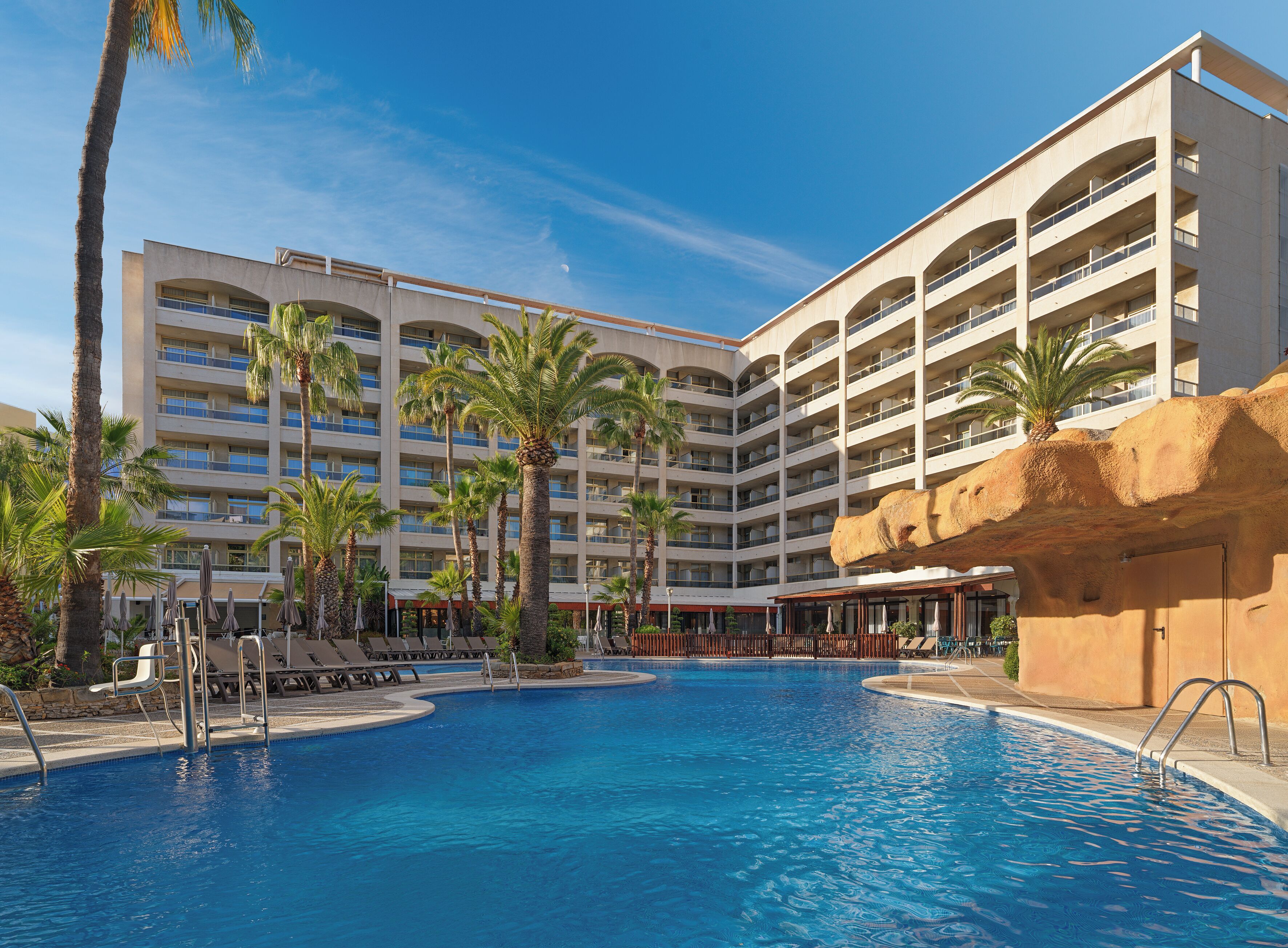Un hotel con una piscina grande, palmeras y un edificio moderno con varios balcones.