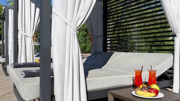 A sunlit outdoor seating area with a cushioned lounge chair, a table with fruit and drinks, and a backdrop of greenery and a metal fence.