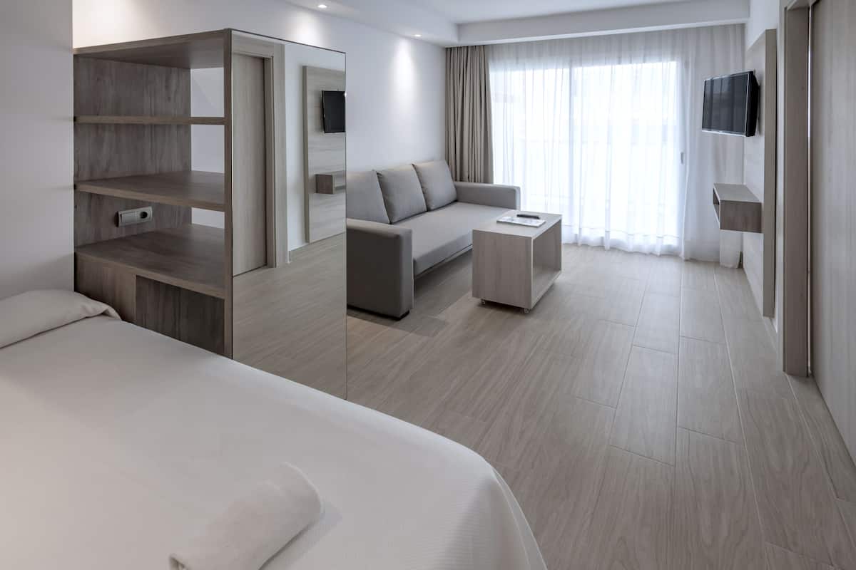 A modern hotel room with a large bed, a sofa, a coffee table, a TV mounted on the wall, and a large window with curtains.