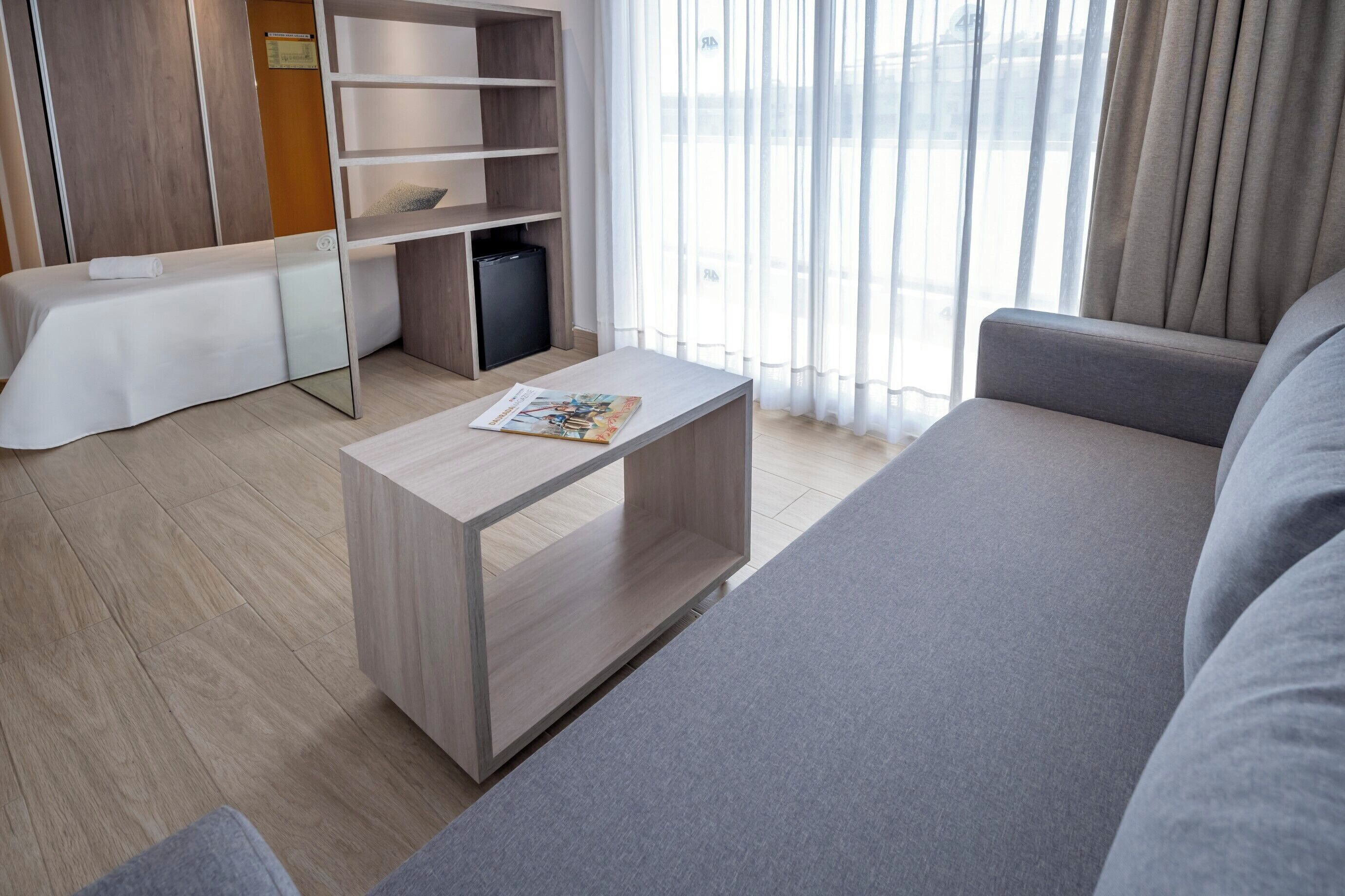 A modern hotel room with a grey sofa, a wooden coffee table with a magazine, a small white table, and a wardrobe.