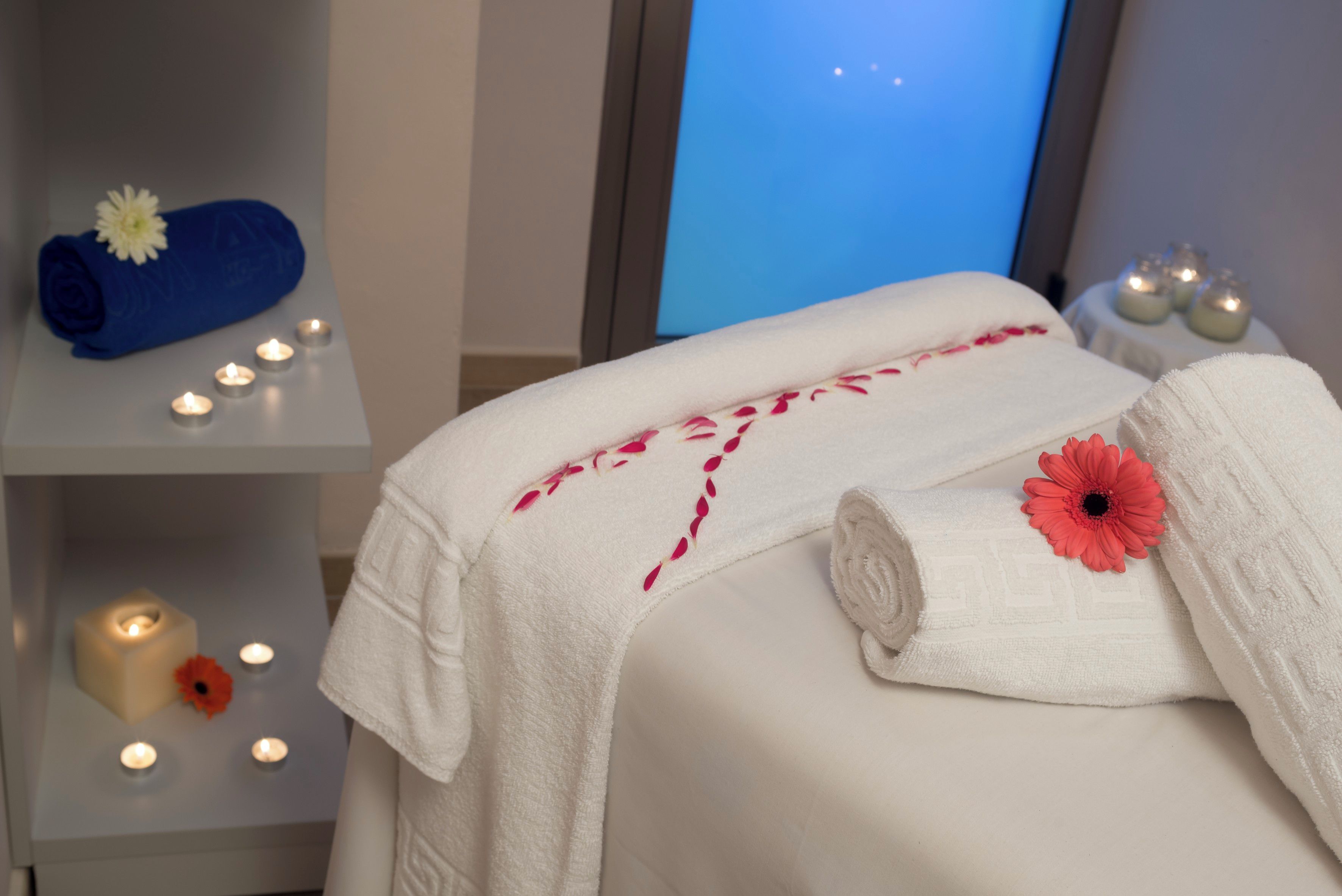Una habitación ordenada con una cama, una toalla azul, una toalla blanca con un detalle floral y varias velas.