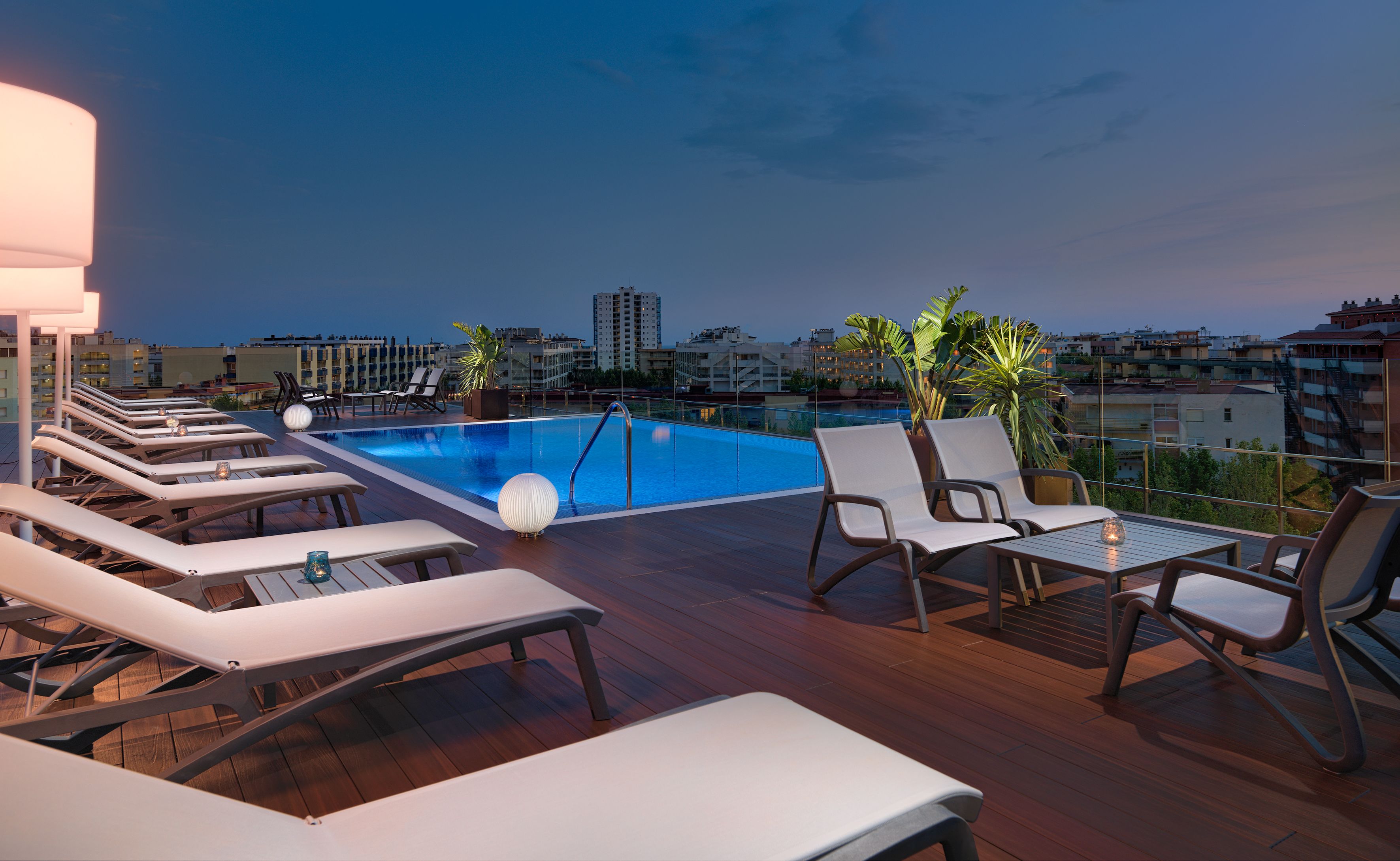 A rooftop pool area with lounge chairs and tables, surrounded by city buildings.