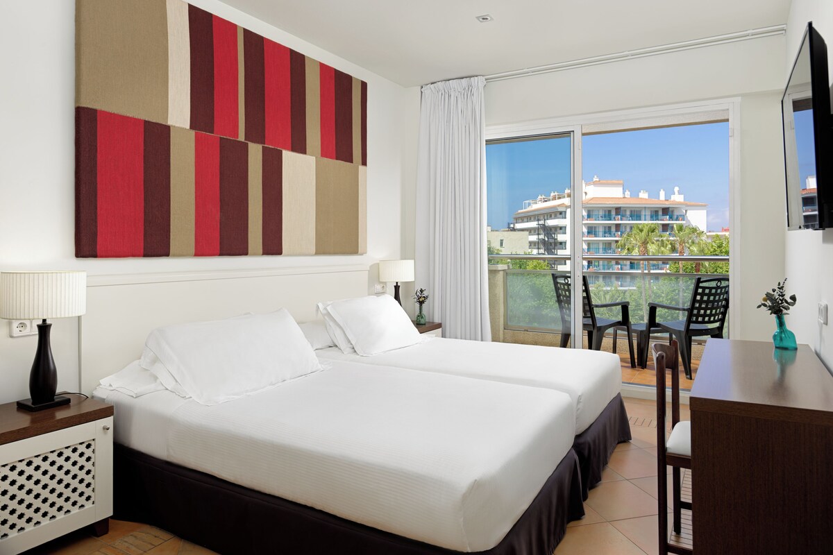 Habitación de hotel con dos camas, un balcón que ofrece vistas a edificios y un escritorio con una silla.