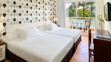 Habitación de hotel con dos camas, un balcón con sillas, televisión y vistas a palmeras.