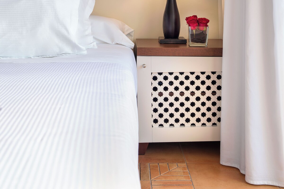 Una cama bien hecha con sábanas blancas, una mesita de noche con una lámpara y un jarrón de rosas rojas, y una pared cubierta con tela a rayas.