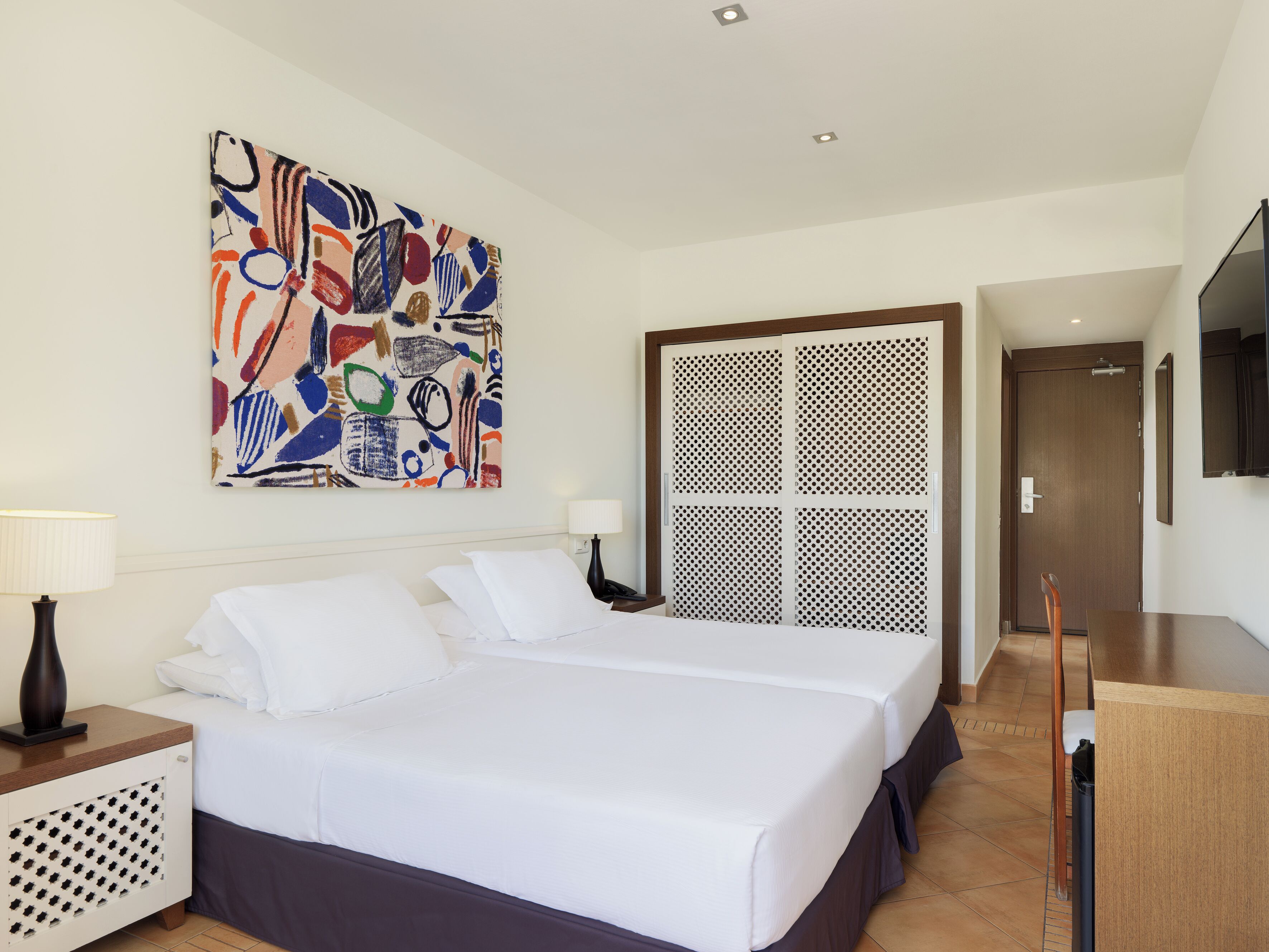 A hotel room with a large bed, a desk, a TV, a painting, and a door.