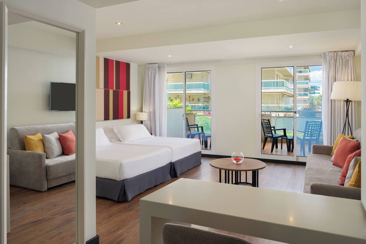 A hotel room with a bed, a sofa, a table, and a balcony with chairs.