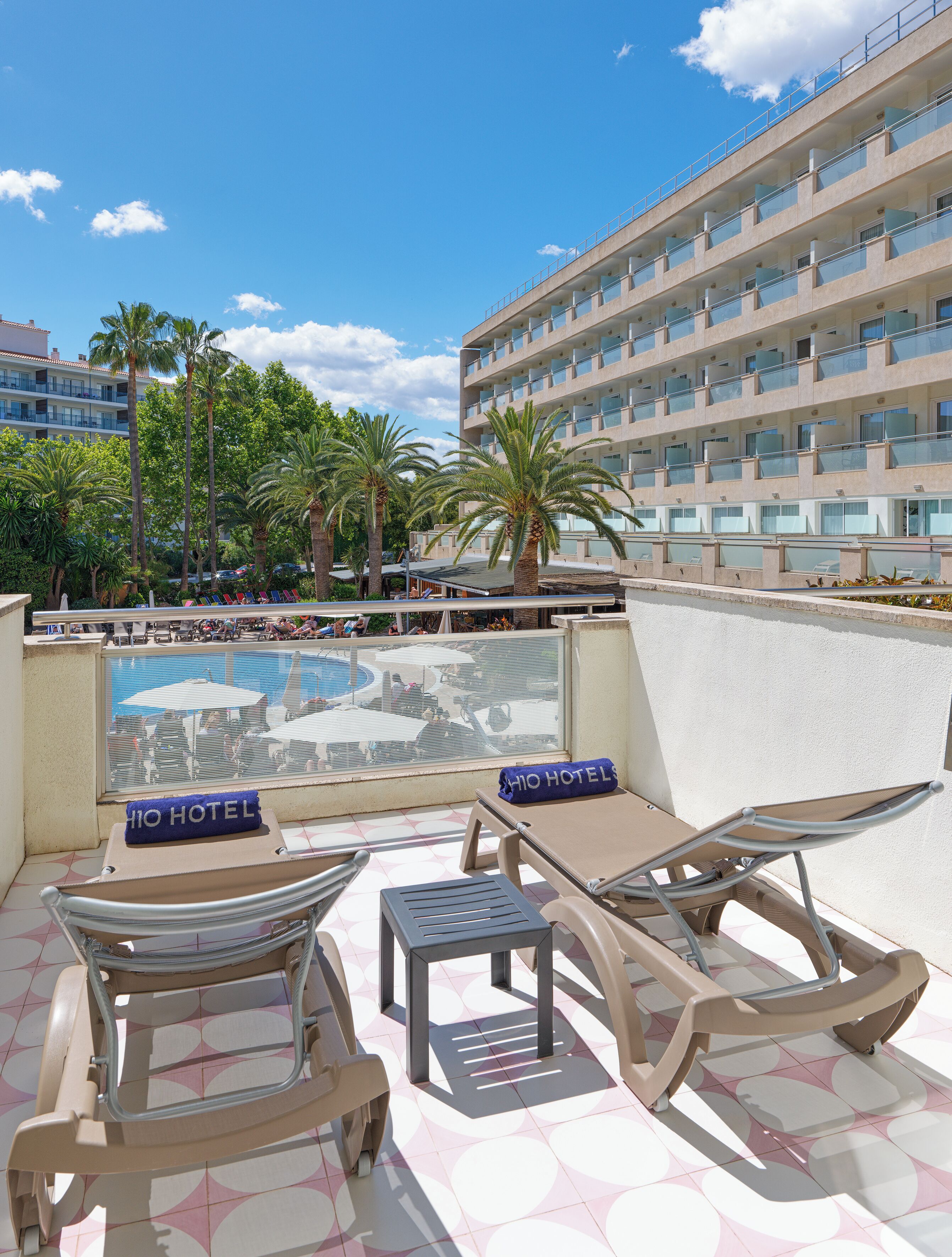 Zona de piscina con sillones de descanso, una mesa pequeña y vistas al edificio del hotel y palmeras.