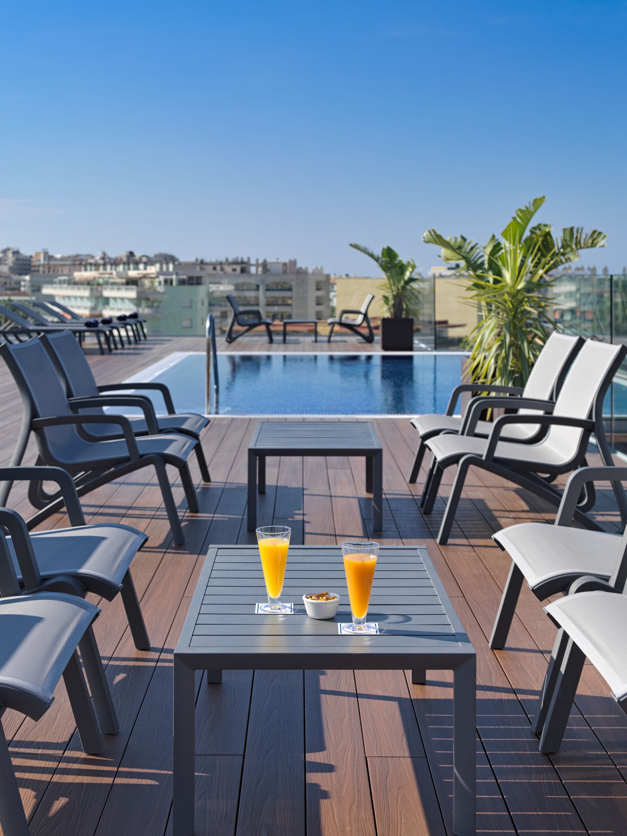 A rooftop pool area with lounge chairs, a table with two glasses of orange juice, and a view of the city skyline.