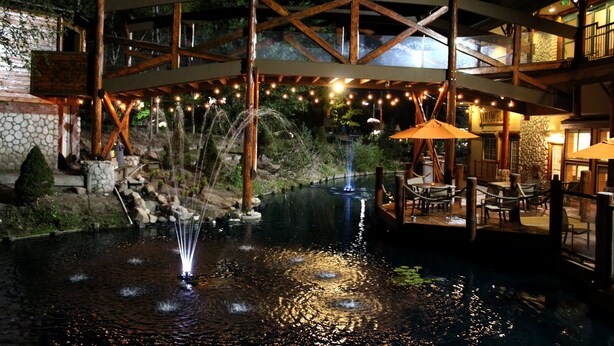 Zona de piscina al aire libre de noche con un puente de madera, luces colgantes y una fuente.
