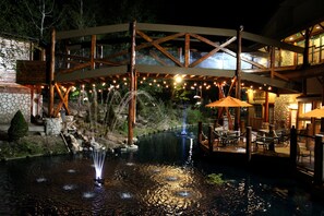 Zona de piscina al aire libre de noche con un puente de madera, luces colgantes y una fuente.