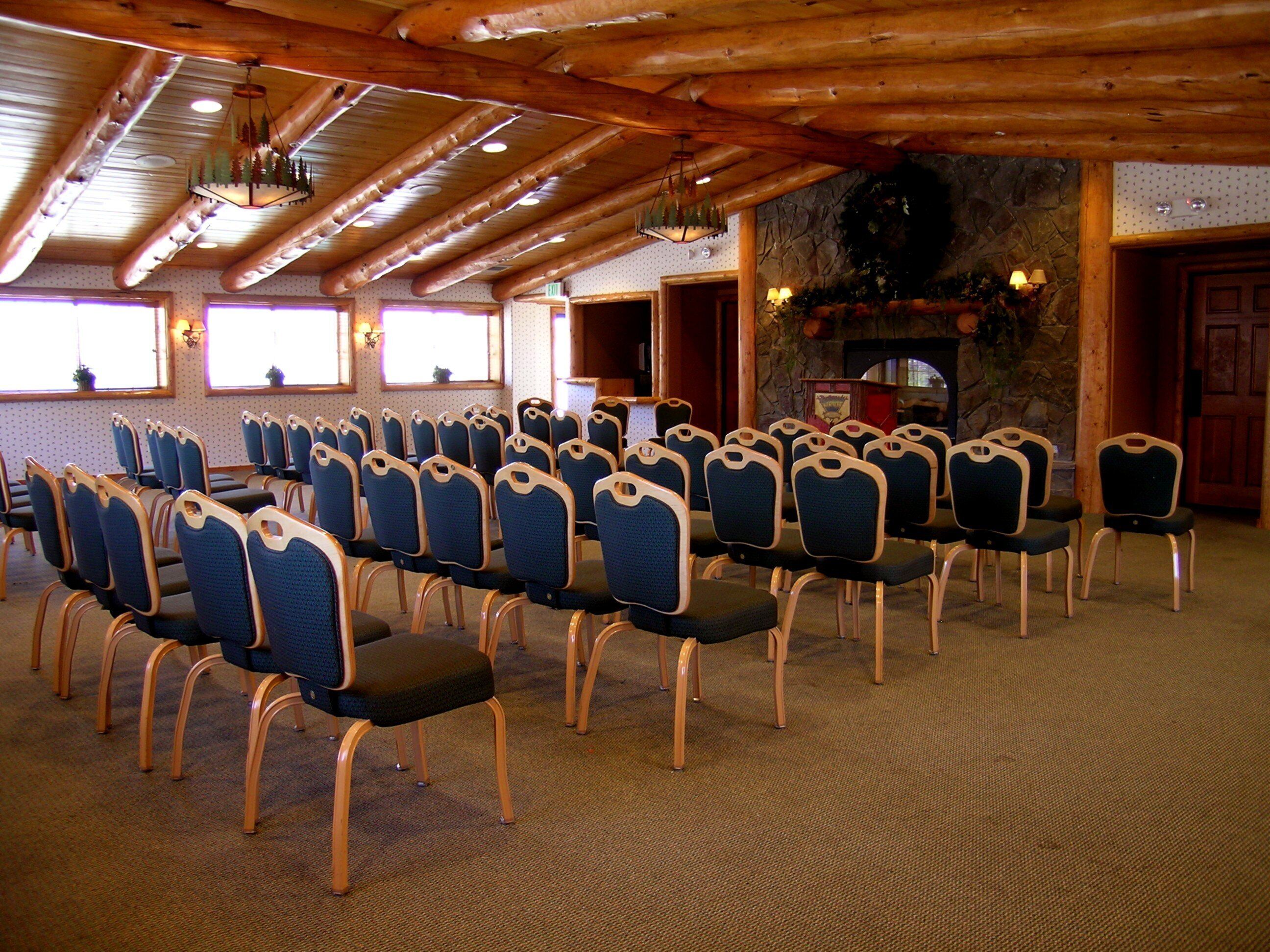 Uma sala de conferências com fileiras de cadeiras, vigas de madeira no teto e uma parede de pedra com lareira.