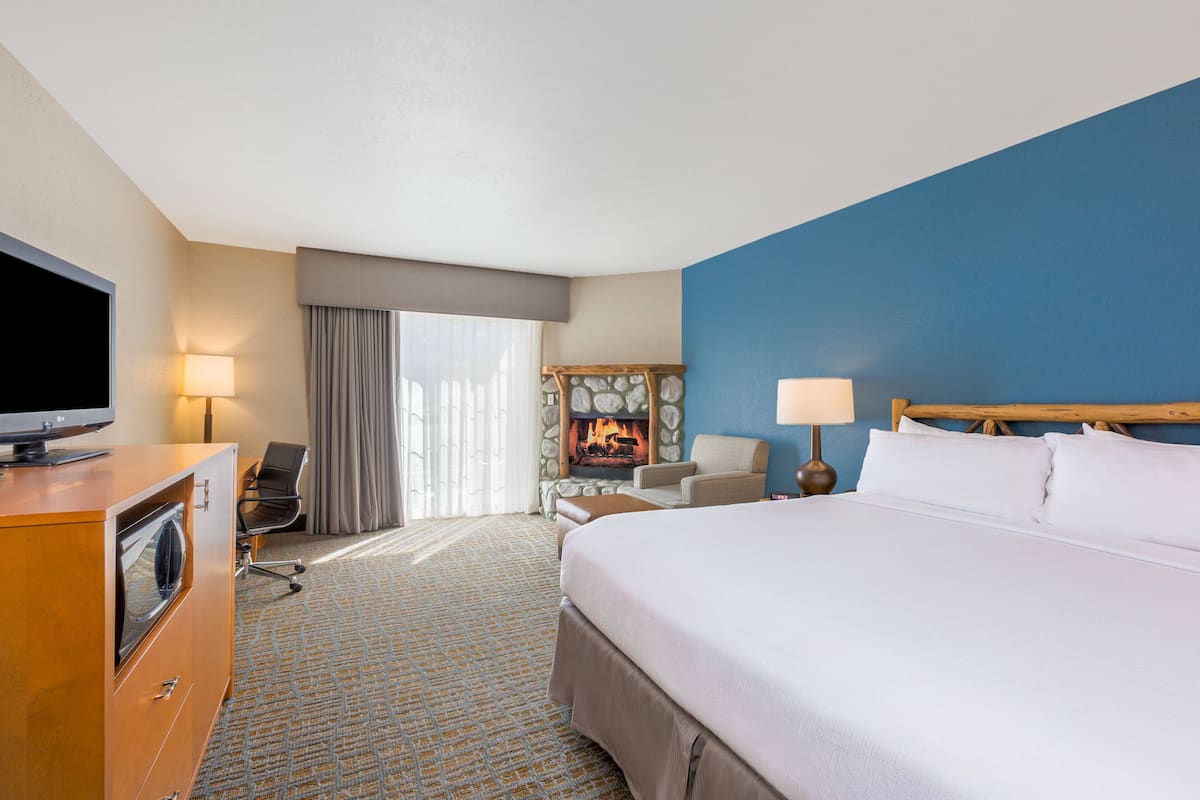 A hotel room with a large bed, a television on a wooden dresser, a desk with a chair, a fireplace, and a window with curtains.