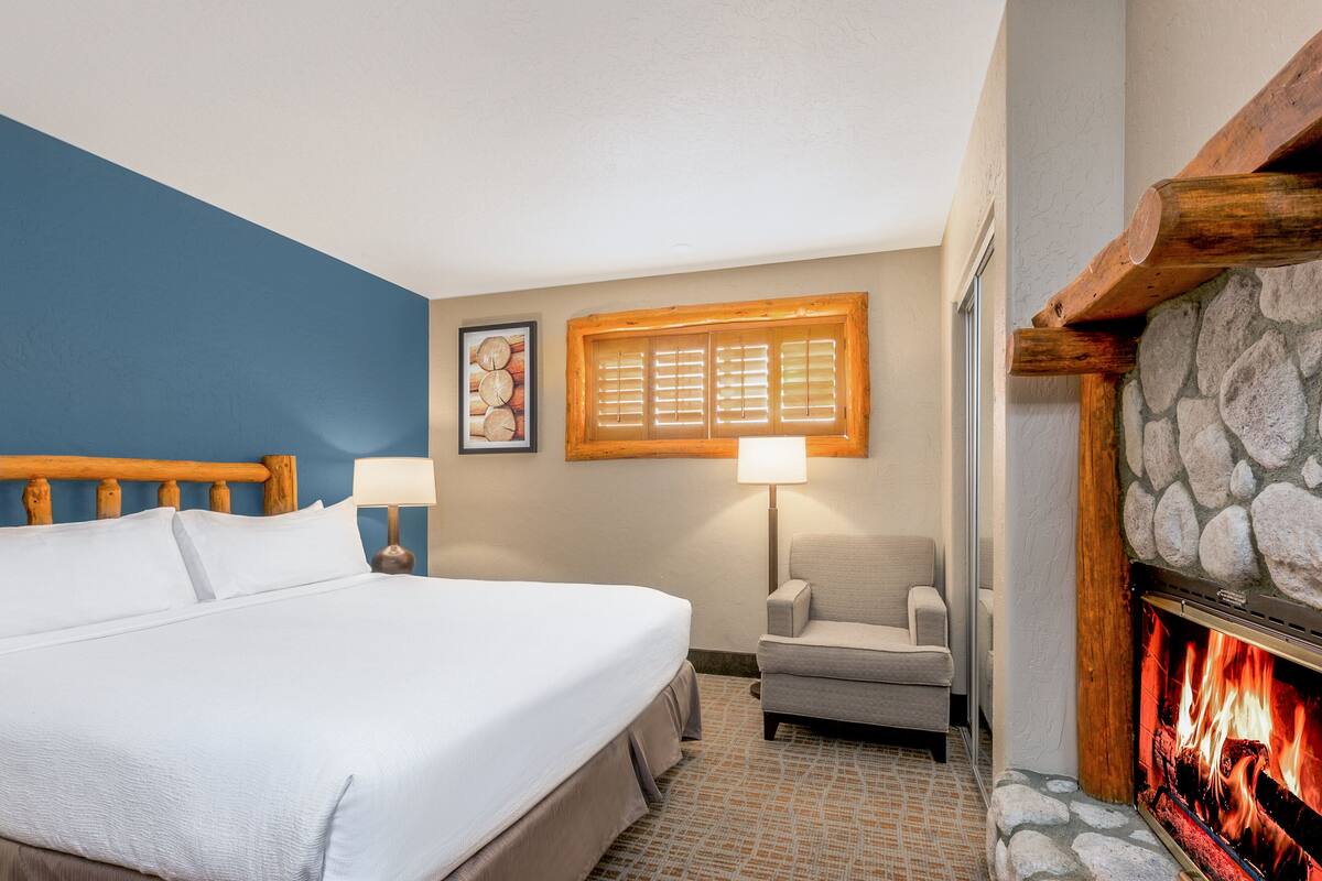 A hotel room with a bed, a chair, a fireplace, a lamp, and a wall art piece.