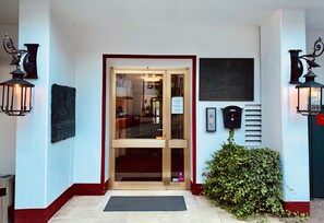 Entrada de hotel com porta de vidro, moldura vermelha e duas lanternas fixadas na parede.