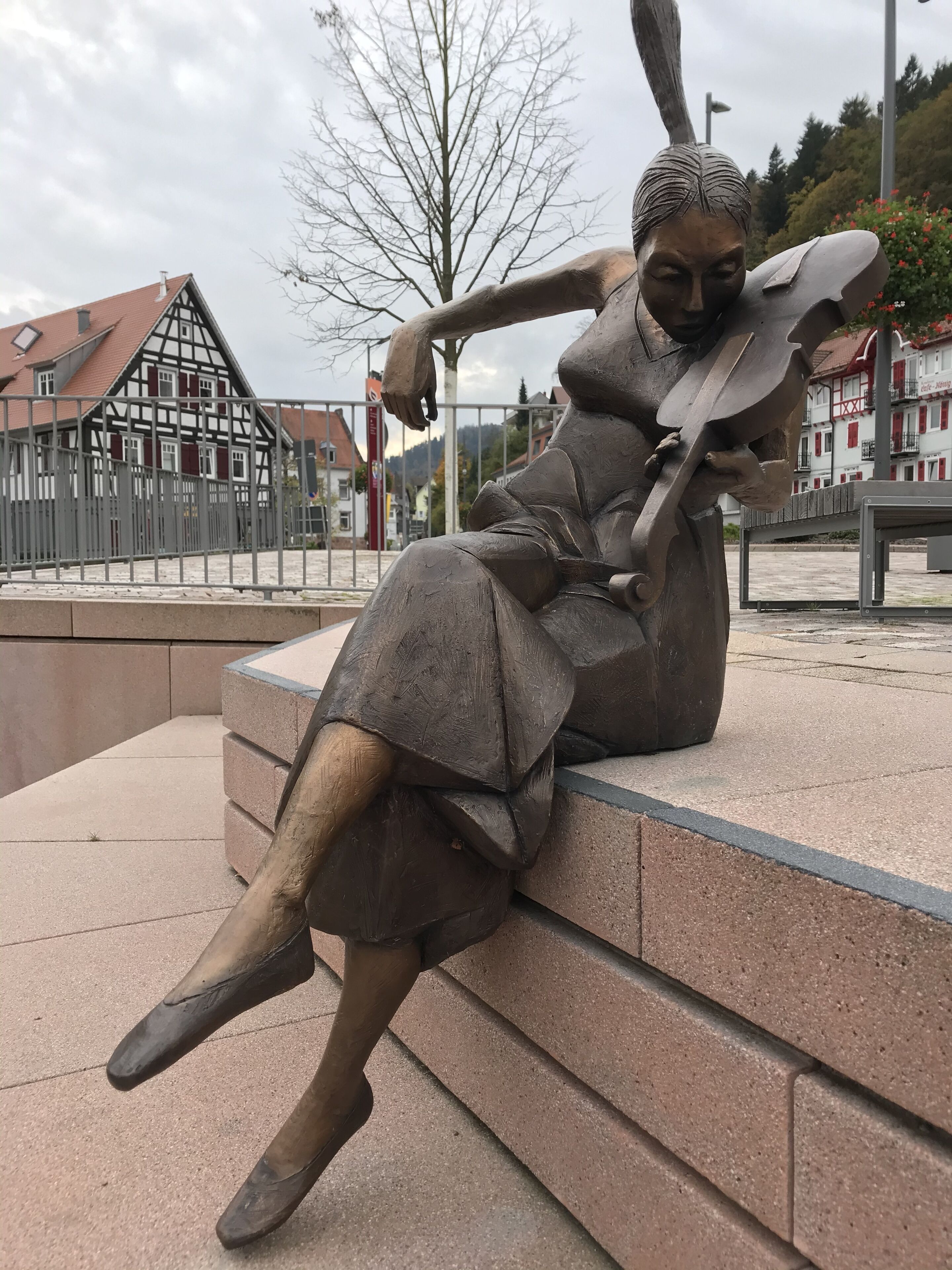 Une statue représentant une figure assise, tenant un livre, avec en arrière-plan des bâtiments traditionnels et un ciel dégagé.